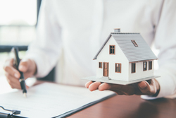 Small model of a home in hand while someone also has a pen in other hand